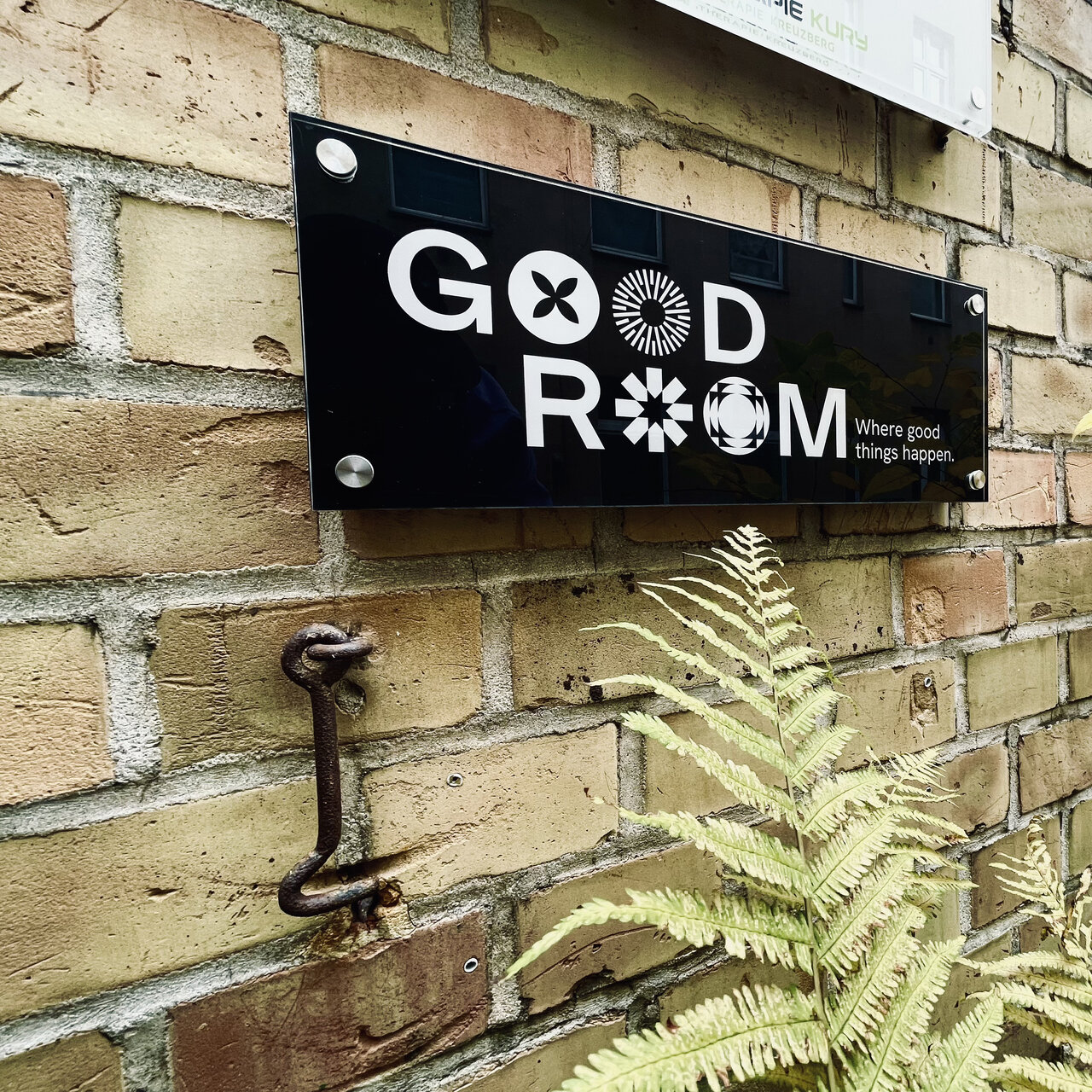 Schwarzes Schild mit "GOOD ROOM" Logo an einer beigen Ziegelwand, mit einer gr&uuml;nen Farnpflanze im Vordergrund und einem rostigen Metallhaken an der Wand.