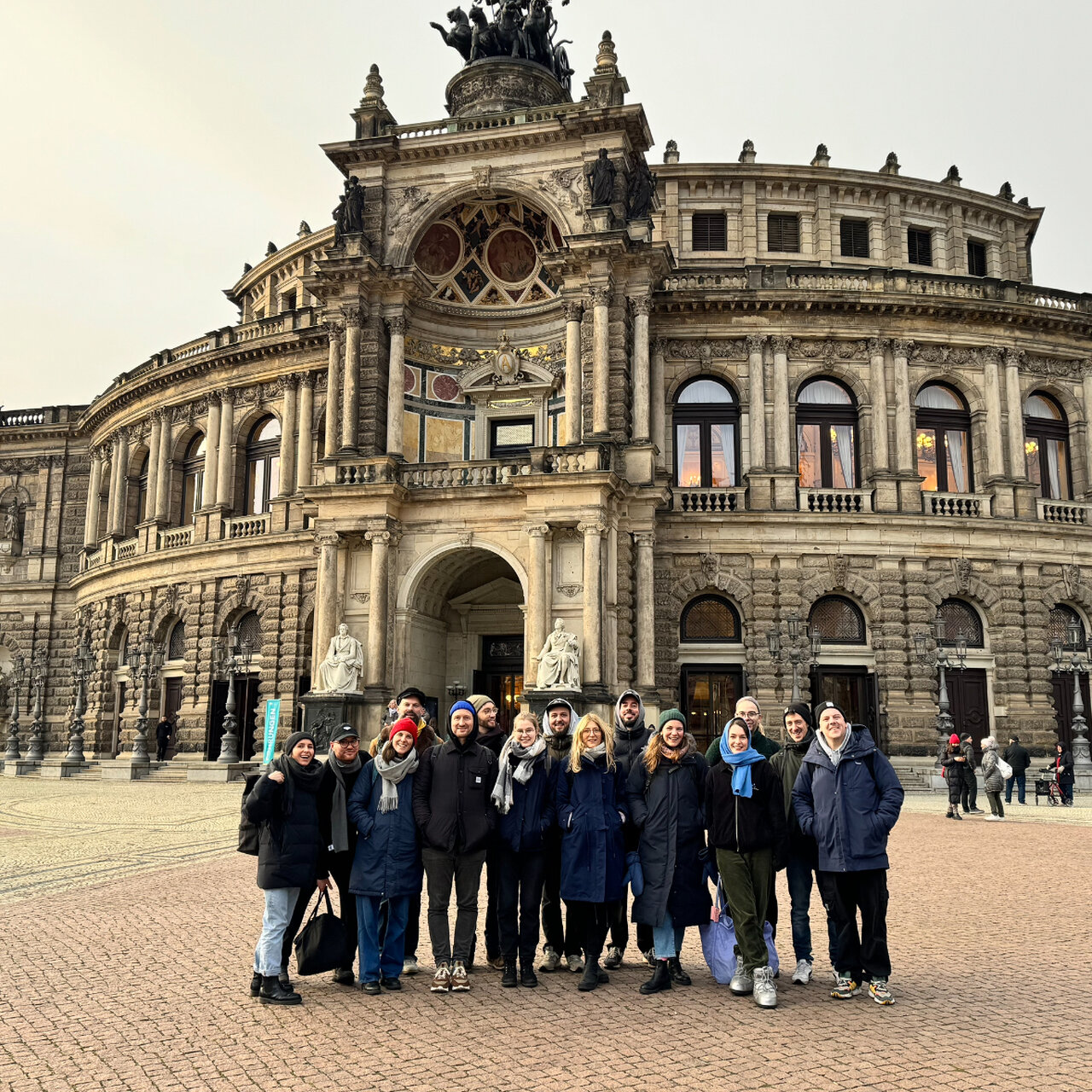 Eine Gruppe von etwa zw&ouml;lf Mitarbeitenden von Studio Good steht in warmen Winterjacken vor dem historischen Semperoper-Geb&auml;ude in Dresden, einem beeindruckenden barocken Opernhaus mit gro&szlig;er Kuppel und verzierter Fassade.