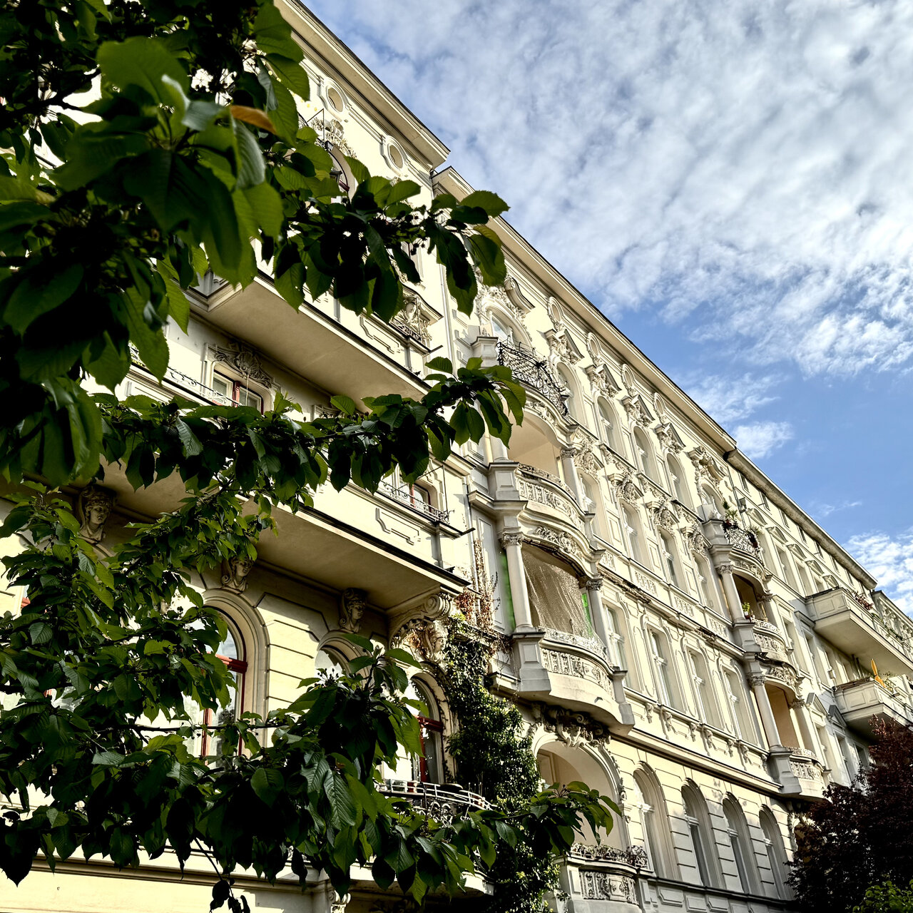 Historische Altbaufassade in Berlin mit Stuck und Verzierungen, wei&szlig;-gelb gestrichen, davor gr&uuml;ne Baumzweige vor blauem Wolkenhimmel. Klassische Architektur mit Erkerfenstern und Balkonen, typisch f&uuml;r das Berliner Stadtbild.