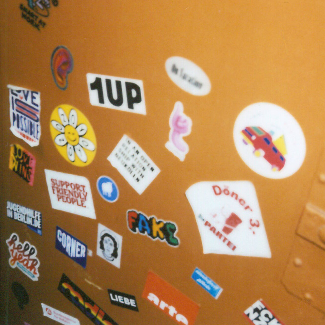 Gelbe Wand mit zahlreichen bunten Stickern und Aufklebern verschiedener Designs, darunter Logos, Grafiken und Schriftz&uuml;ge wie '1UP' und andere kreative Motive.