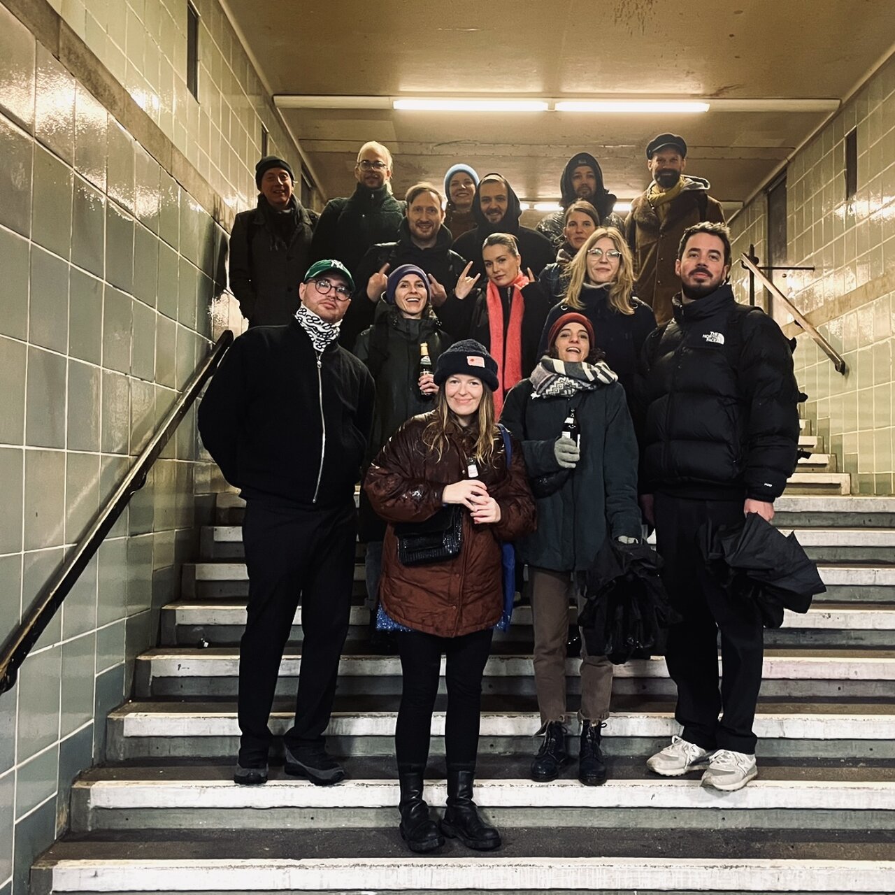 Eine Gruppe von etwa 15 Personen steht auf einer Treppe in einem Treppenhaus mit gefliesten W&auml;nden. Alle tragen Winterkleidung in dunklen Farben und stehen eng beieinander f&uuml;r ein Gruppenfoto.