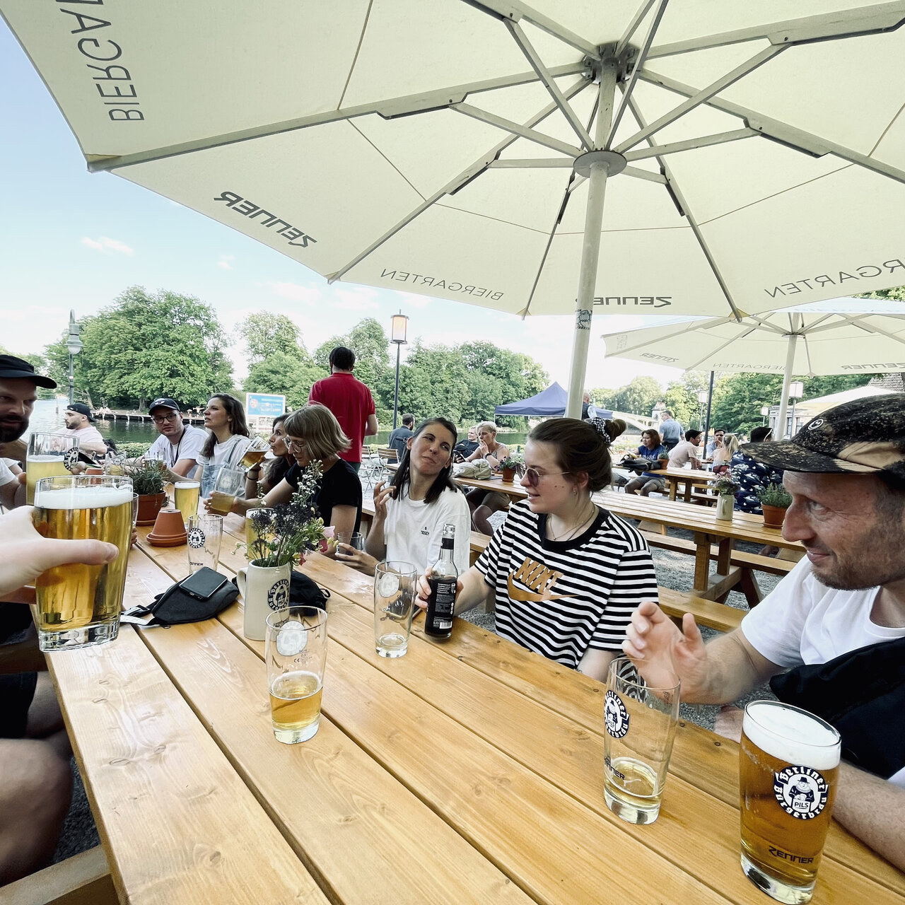 Eine Gruppe von Menschen sitzt an Holztischen unter einem gro&szlig;en Sonnenschirm und genie&szlig;t Bier und Getr&auml;nke in entspannter Atmosph&auml;re an einem sonnigen Tag.