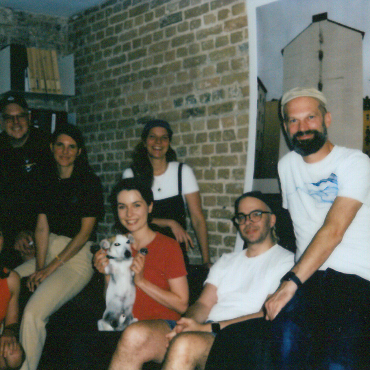 Sechs Personen in einem Ziegelsteinraum, sitzen und stehen zusammen, eine Person h&auml;lt einen Hund. Eine entspannte B&uuml;roatmosph&auml;re bei Studio Good, die Teamzusammenhalt und Kreativit&auml;t ausstrahlt.