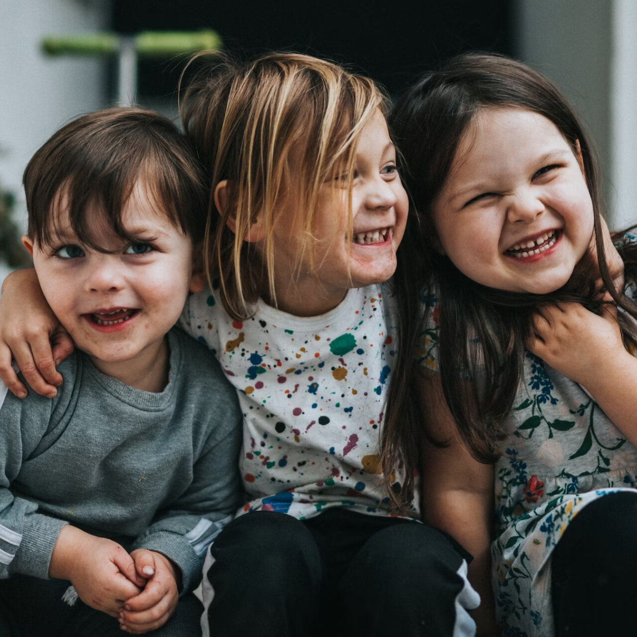 Drei lachende Kinder sitzen nebeneinander, der Mittleren umarmt die anderen beiden. Sie tragen lockere Kleidung und strahlen pure Freude und Verbundenheit aus.