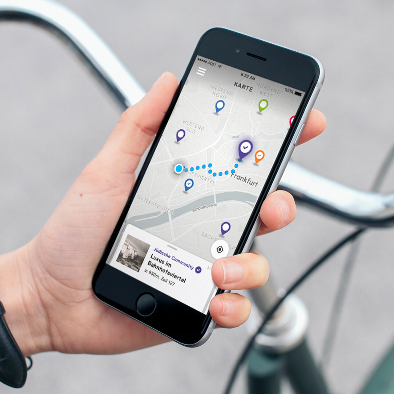 Eine Hand h&auml;lt ein Smartphone mit einer Navigations-App, die einen Stadtplan mit blauen und gr&uuml;nen Markierungspunkten zeigt. Die App scheint eine Routen- oder Standortverfolgung auf einem Hintergrund mit einem Fahrradlenker zu pr&auml;sentieren.