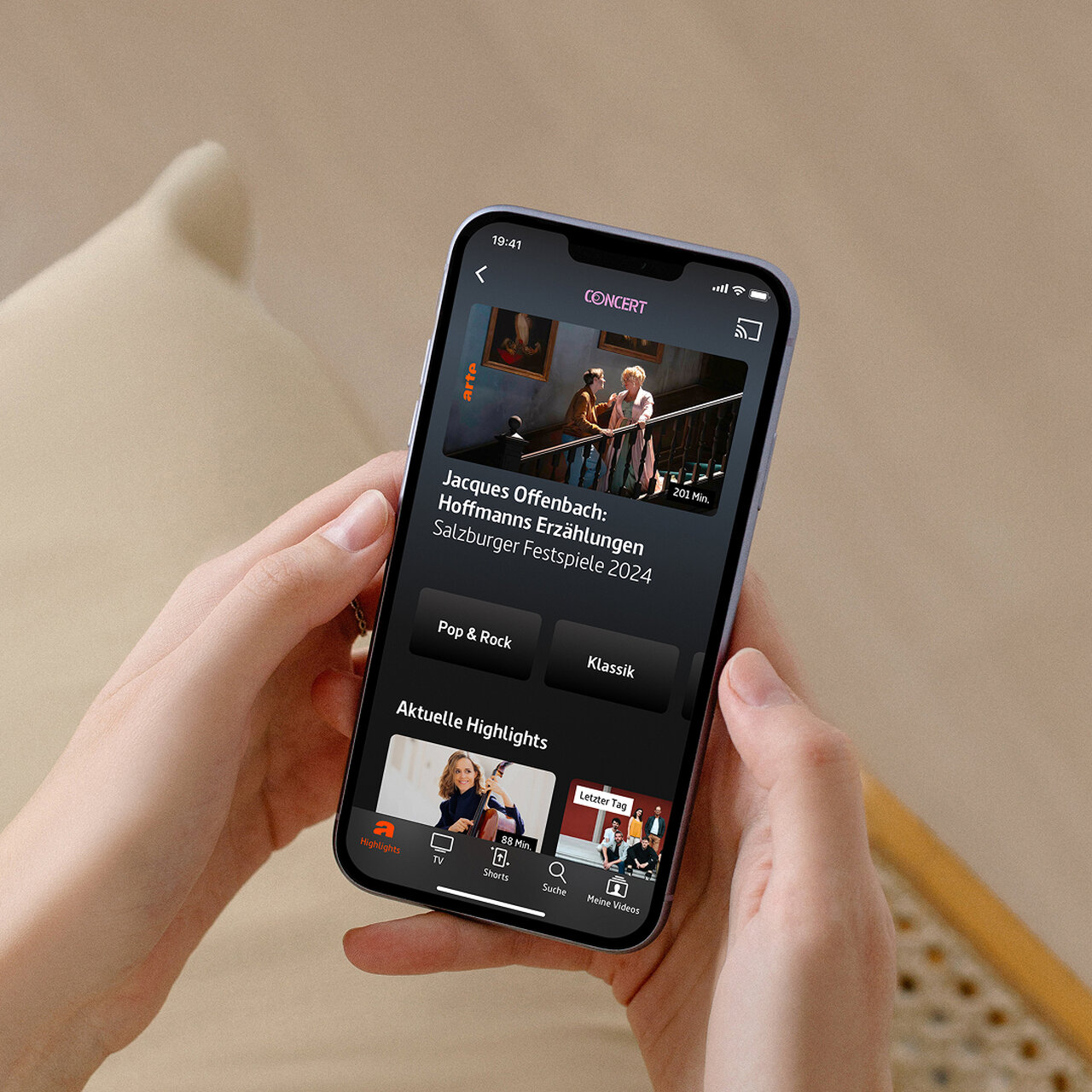 Eine Hand h&auml;lt ein Smartphone mit einer Musik- oder Konzert-App, die ein Konzertprogramm und Highlights auf dem Bildschirm zeigt, mit einem Bild von zwei Personen auf einer Treppe im oberen Bereich.