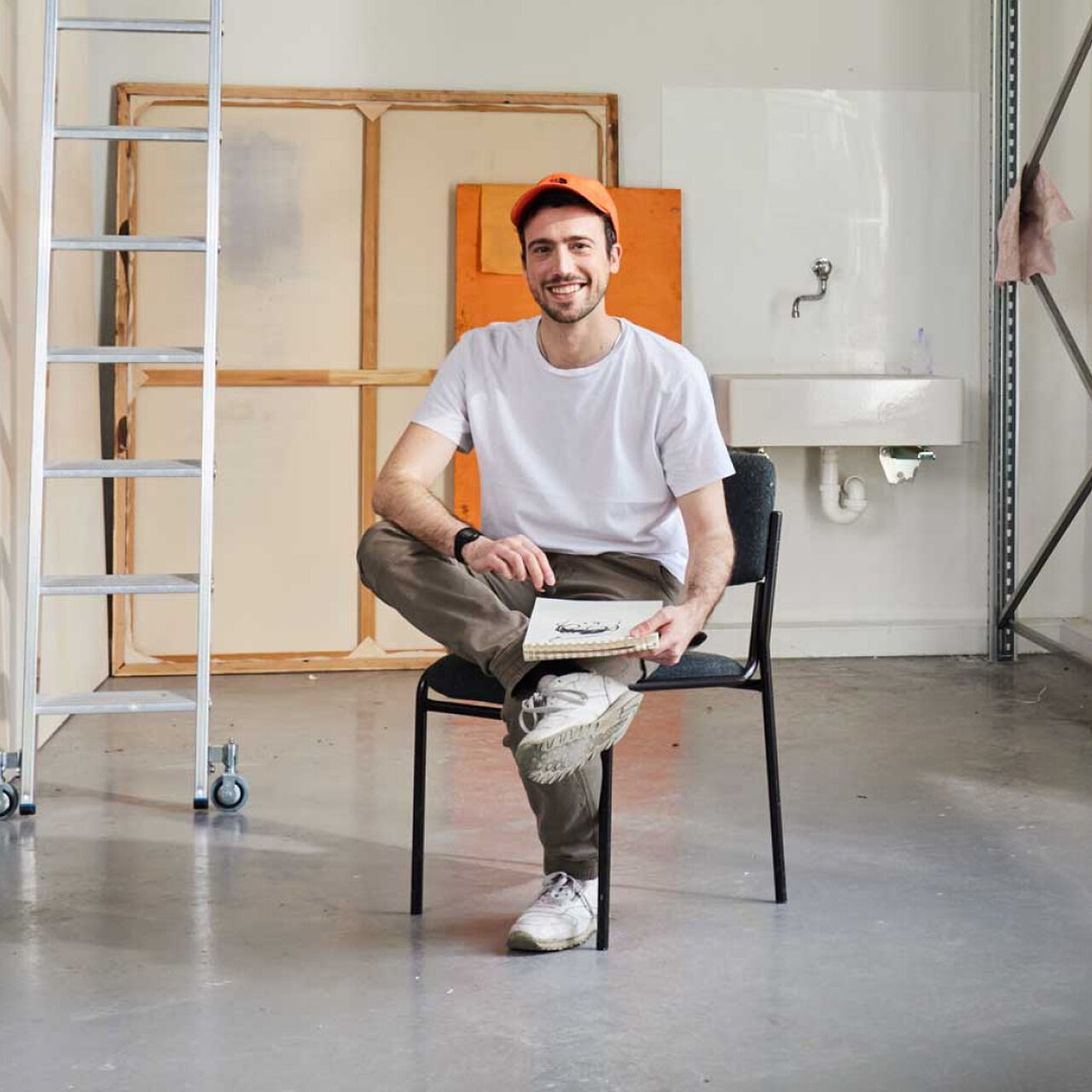 Ein l&auml;chelnder Mann in wei&szlig;em T-Shirt und Baseballkappe sitzt entspannt auf einem schwarzen Stuhl in einem minimalistischen Raum mit Leiter und orangefarbenem Kunstwerk im Hintergrund.
