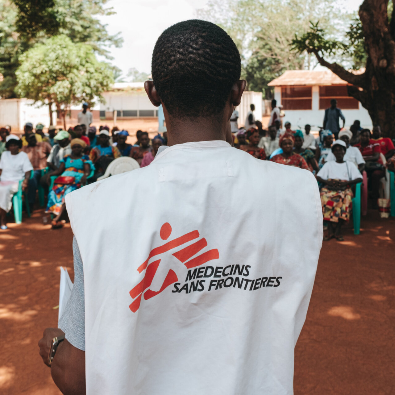 Mitarbeiter von M&eacute;decins Sans Fronti&egrave;res (&Auml;rzte ohne Grenzen) von hinten, gekleidet in einer wei&szlig;en Weste mit dem roten Organisationslogo, vor einer Menschenmenge auf einem roten Lehmplatz in einer l&auml;ndlichen Umgebung mit B&auml;umen und Geb&auml;uden im Hintergrund.