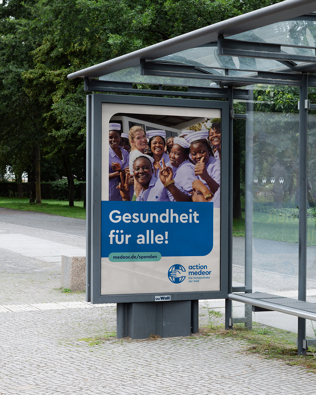 Mehrere Menschen in wei&szlig;en Hemden l&auml;cheln und gestikulieren an einem Bushaltestellenschild mit dem Slogan "Gesundheit f&uuml;r alle!" von Action Medecor, das die Bedeutung globaler Gesundheitsversorgung in Krisensituationen unterstreicht.