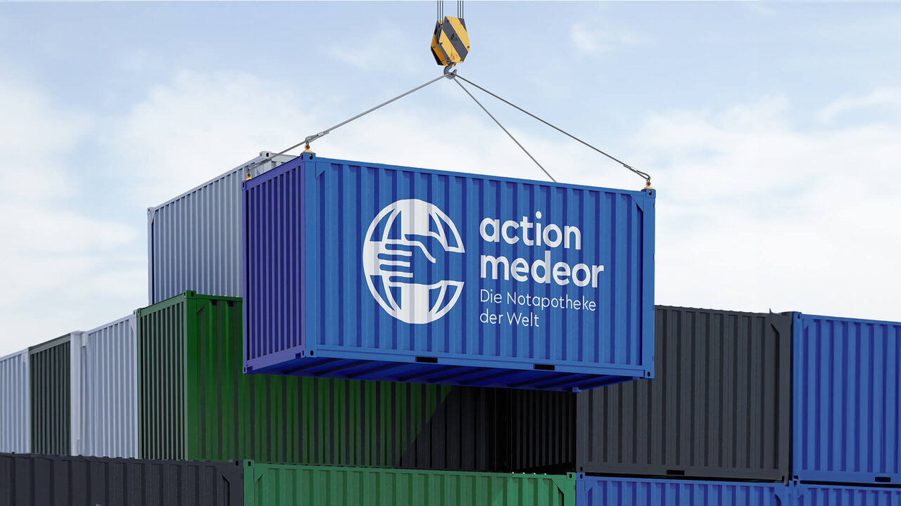 Ein blauer Seecontainer mit dem Logo von action medeor, der an einem Kran h&auml;ngt und zwischen anderen Containern in verschiedenen Farben positioniert ist, vor einem hellen Himmel.