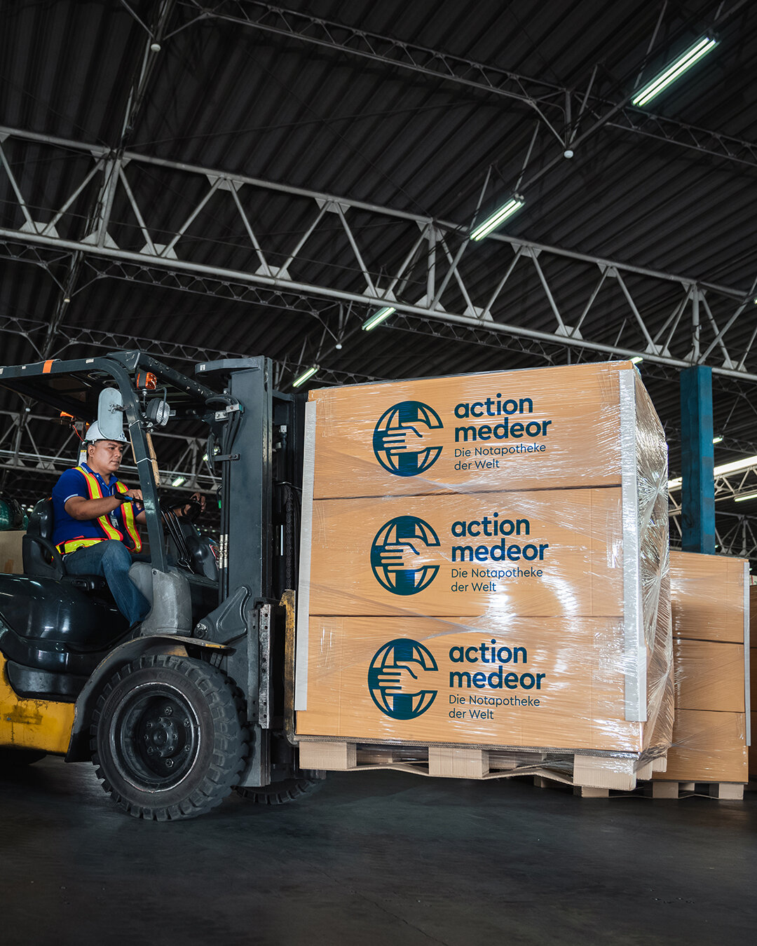 Gabelstapler transportiert drei Holzpaletten mit action medeor-Logo in einem Industrielager, beladen mit medizinischen Hilfsg&uuml;tern und verpackt in Plastikfolie.