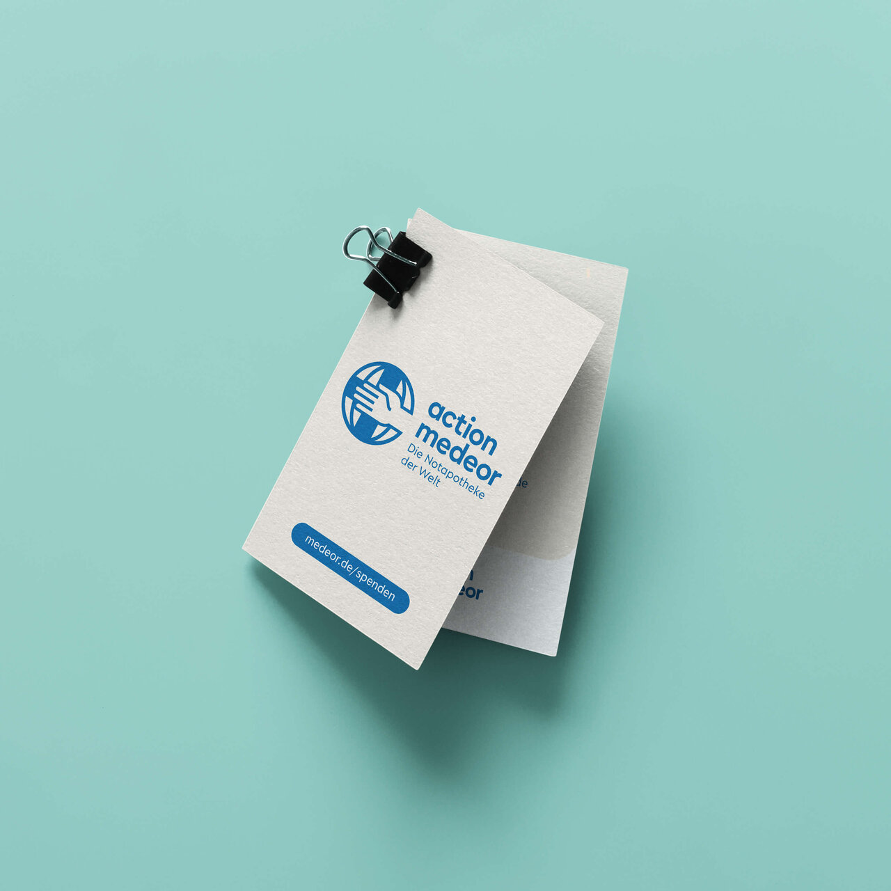 Visitenkarte von action medeor mit blauem Logo auf cremefarbenem Papier, befestigt mit einer schwarzen B&uuml;roklammer auf mintgr&uuml;nem Hintergrund.