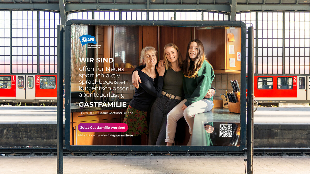Drei Frauen verschiedener Generationen stehen eng beieinander in einem Plakatwerbungsrahmen an einer U-Bahn-Station. Sie tragen moderne Kleidung in dunklen Farbt&ouml;nen und l&auml;cheln selbstbewusst in die Kamera. Das Plakat pr&auml;sentiert eine Kampagne f&uuml;r Gastfamilien mit dem Slogan "Wir sind sportlich aktiv, sprachbegeistert und offen".