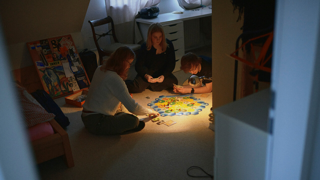 Drei Personen sitzen im ged&auml;mpften Licht um ein gro&szlig;es, beleuchtetes Puzzle, das auf dem Boden ausgebreitet ist. Im Hintergrund sind ein B&uuml;cherregal und M&ouml;bel zu sehen, w&auml;hrend die Gruppe konzentriert zusammen am Puzzle arbeitet.