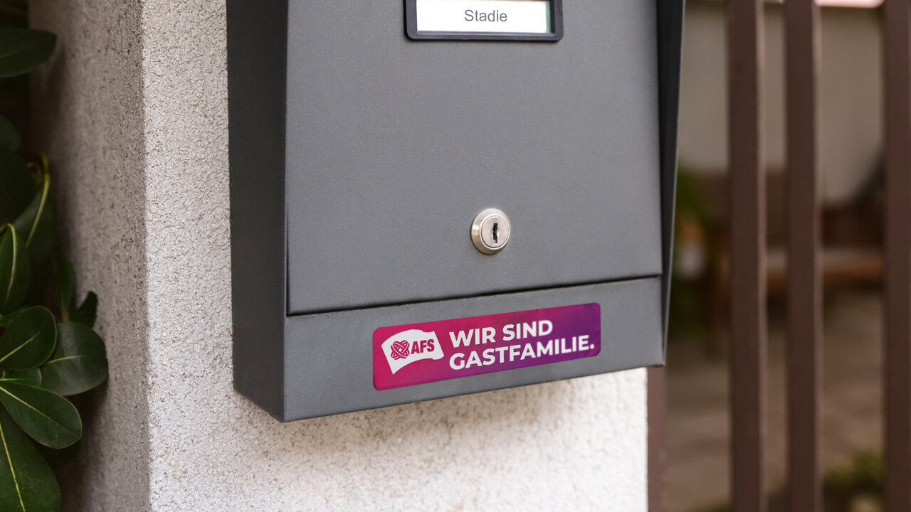 Grauer Briefkasten mit einem pinkfarbenen Aufkleber der DARS-Gastfamilie. Der Aufkleber zeigt den Text "WIR SIND GASTFAMILIE." vor einem wei&szlig;en Hintergrund.