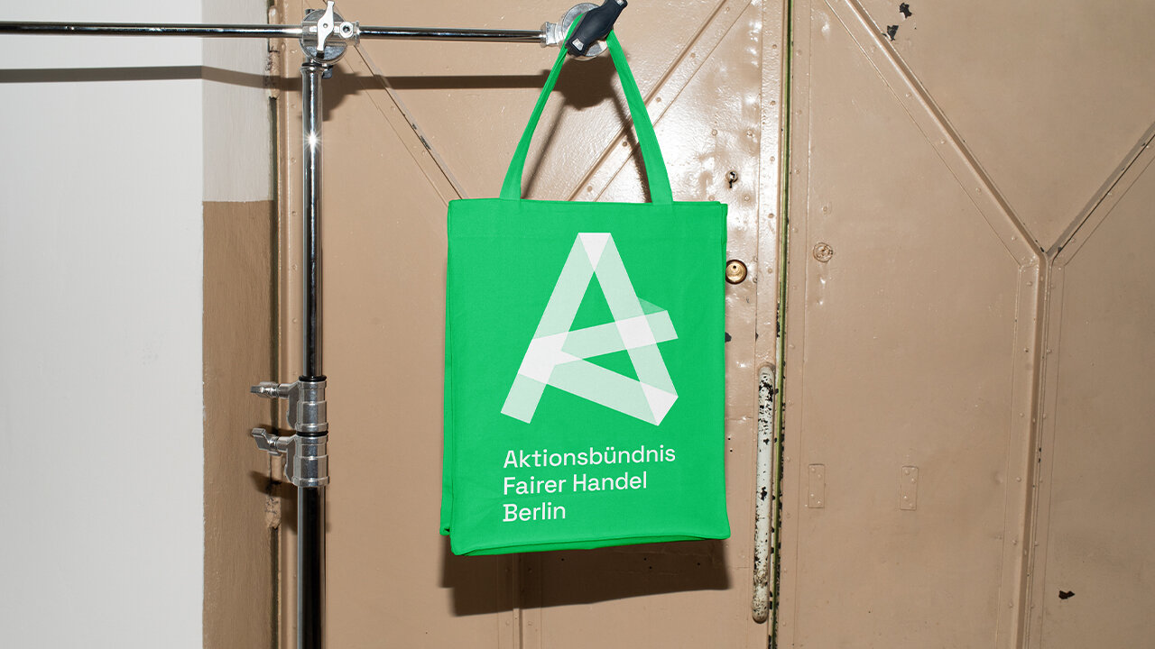 Gr&uuml;ne Einkaufstasche des Aktionsb&uuml;ndnisses Fairer Handel Berlin, mit wei&szlig;em A-Logo und Schriftzug, h&auml;ngt an einer Metallstange vor einem beigen Hintergrund.
