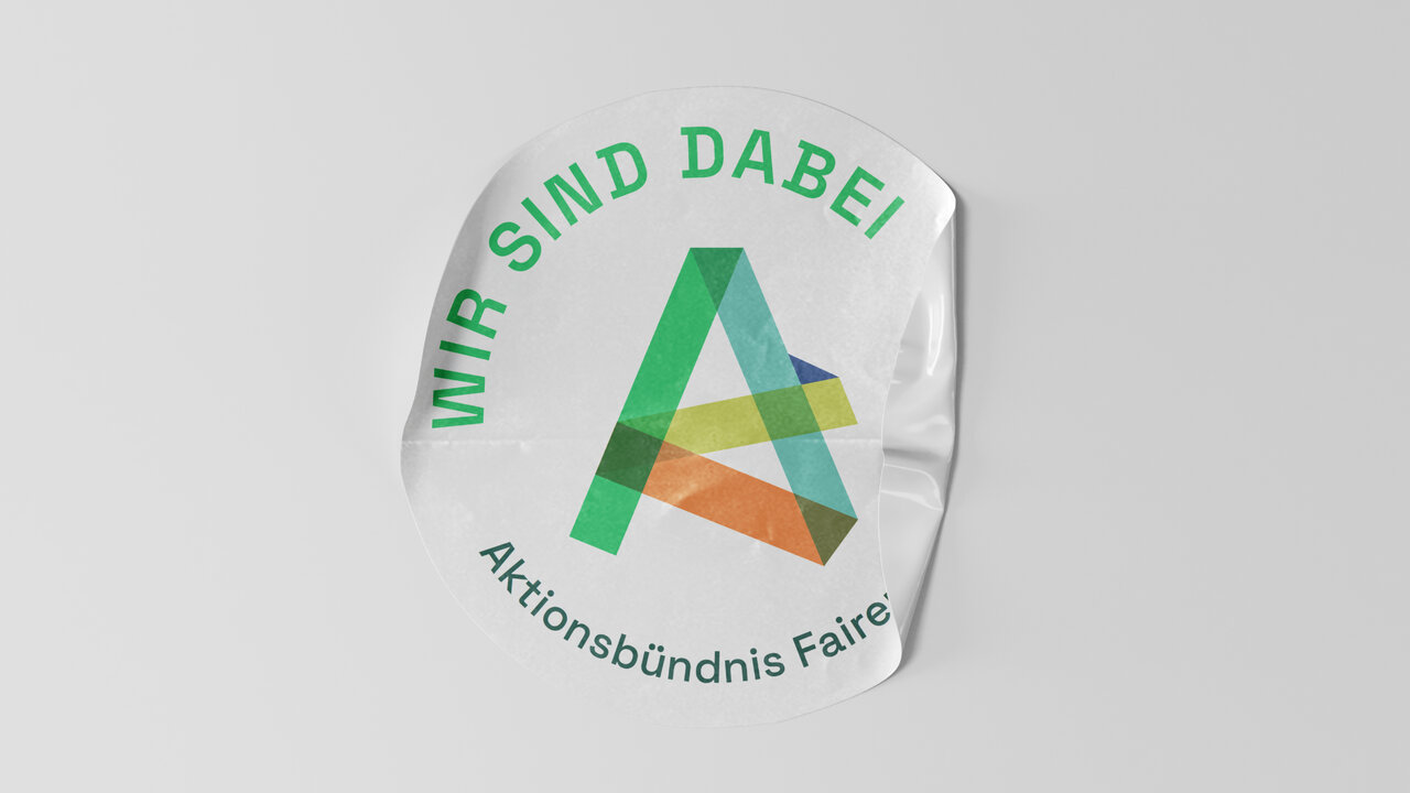 Rundes Sticker-Design des Aktionsb&uuml;ndnisses Fairer Handel in Gr&uuml;n- und Oranget&ouml;nen mit gestapeltem A-Logo und dem Slogan "Wir sind dabei".