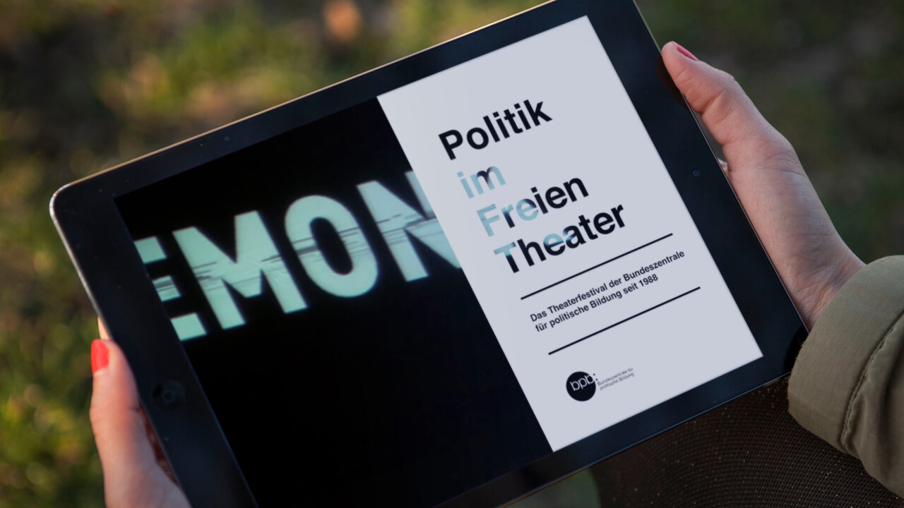 Tablet-Bildschirm zeigt ein digitales Magazin mit dem Titel "Politik im Freien Theater" vor unscharfem Hintergrund, gehalten von einer Hand mit rotem Nagellack.