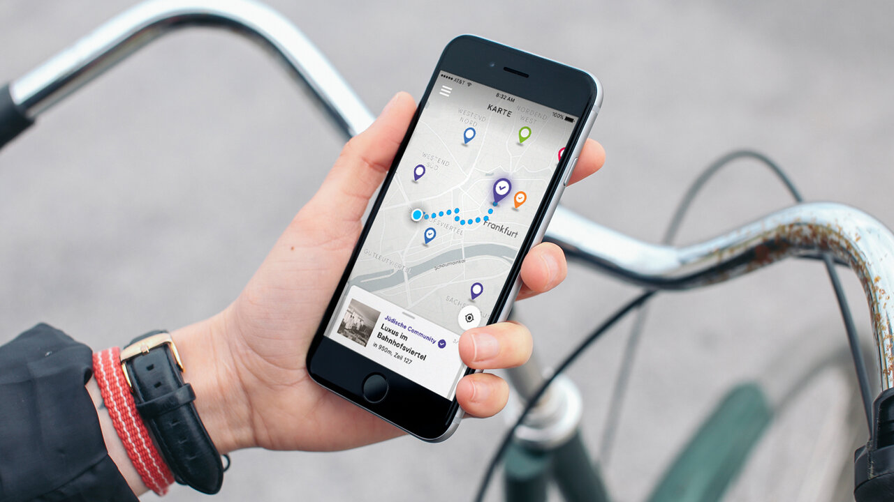 Hand h&auml;lt Smartphone mit ge&ouml;ffneter Navigations-App, das Fahrradlenker im Hintergrund zeigt. Auf dem Bildschirm ist eine interaktive Stadtkarte mit mehreren Markierungspunkten zu sehen, die eine Routenf&uuml;hrung visualisiert.