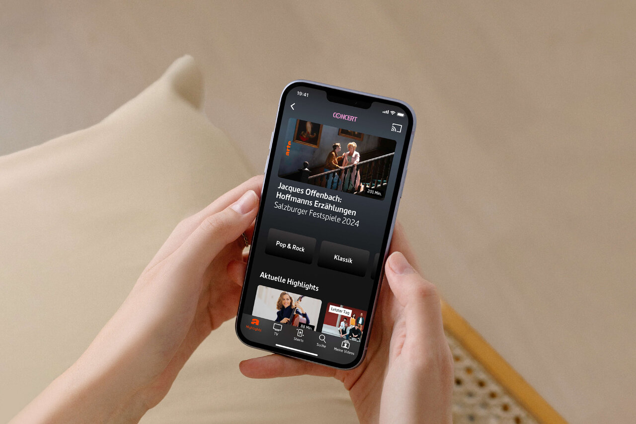 Smartphone-Bildschirm zeigt die ARTE-App mit einem Konzertbild und Programmvorschl&auml;gen, gehalten von einer Hand vor einem beigen Hintergrund.