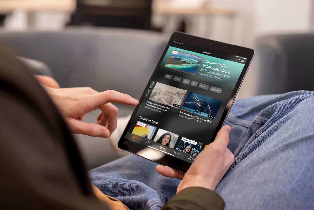 Tablet-Bildschirm zeigt die ARTE-App mit verschiedenen Videoinhalten und Vorschaubildern, w&auml;hrend eine Hand das Ger&auml;t in einer entspannten Sitzposition bedient.