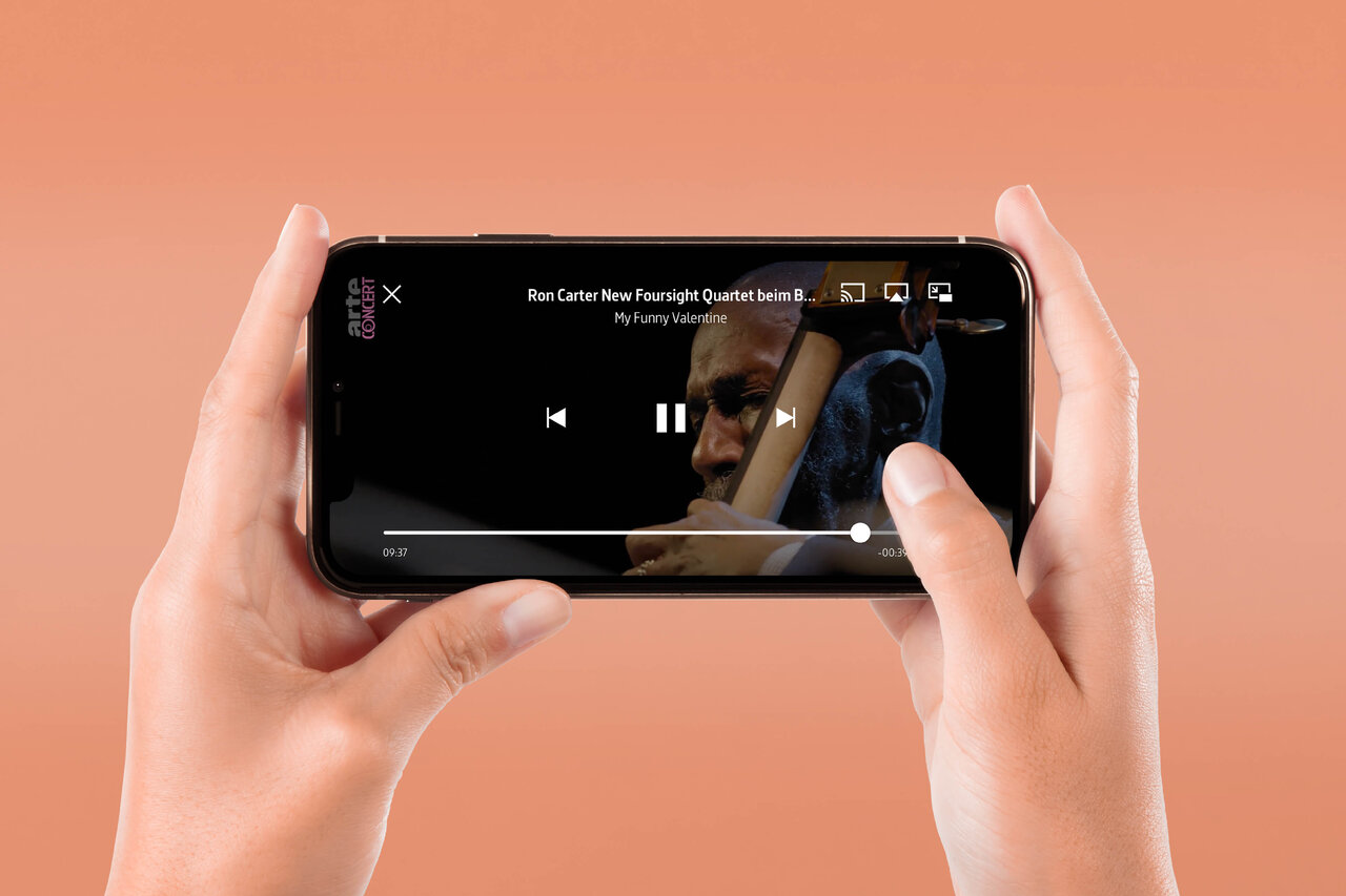 Eine Nahaufnahme von H&auml;nden, die ein Smartphone horizontal halten und einen Videoplayer mit pausiertem Inhalt auf dem Bildschirm zeigen, vor einem pfirsichfarbenen Hintergrund.