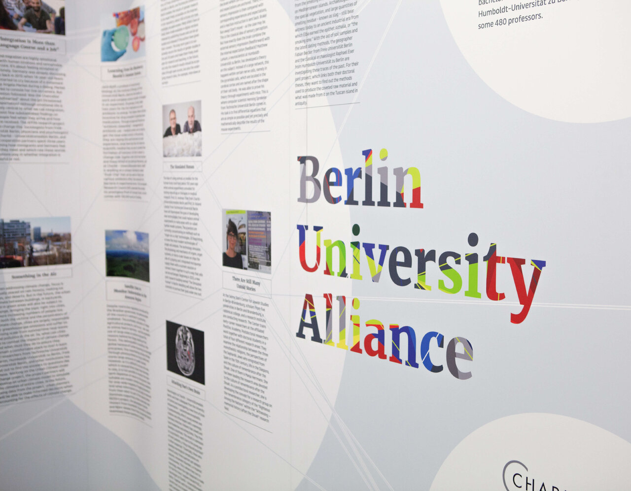 Berlin University Alliance Logo in bunten Buchstaben auf einem wei&szlig;en Ausstellungsstand mit textilem Hintergrund und seitlichen Informationstafeln.