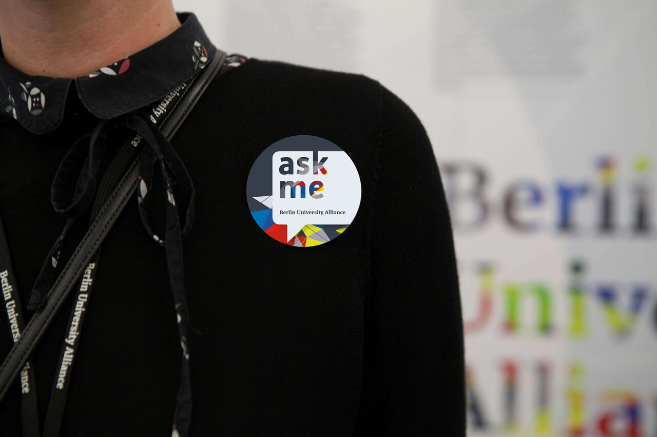 Auf der schwarzen Jacke eines Mitarbeiters ist ein rundes "ask me" Sticker mit bunten grafischen Elementen angebracht, der vor einem unscharfen Hintergrund mit Schriftz&uuml;gen zu sehen ist.