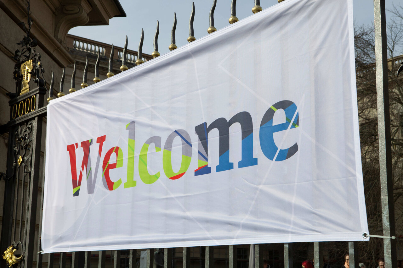 Gro&szlig;e wei&szlig;e Flagge mit buntem "Welcome"-Schriftzug, aufgeh&auml;ngt an einem Zaun vor einem historischen Geb&auml;ude.