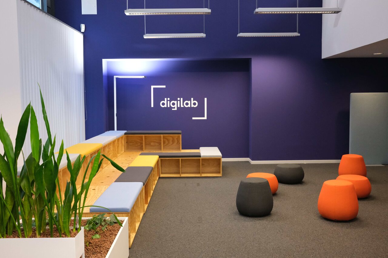 Moderner Wartebereich mit blauer Akzentwand und "Digilab"-Logo, ausgestattet mit Holzsitzgelegenheiten, Gr&uuml;npflanzen, grauen und orangefarbenen Sitzm&ouml;beln auf einem grauen Teppichboden.
