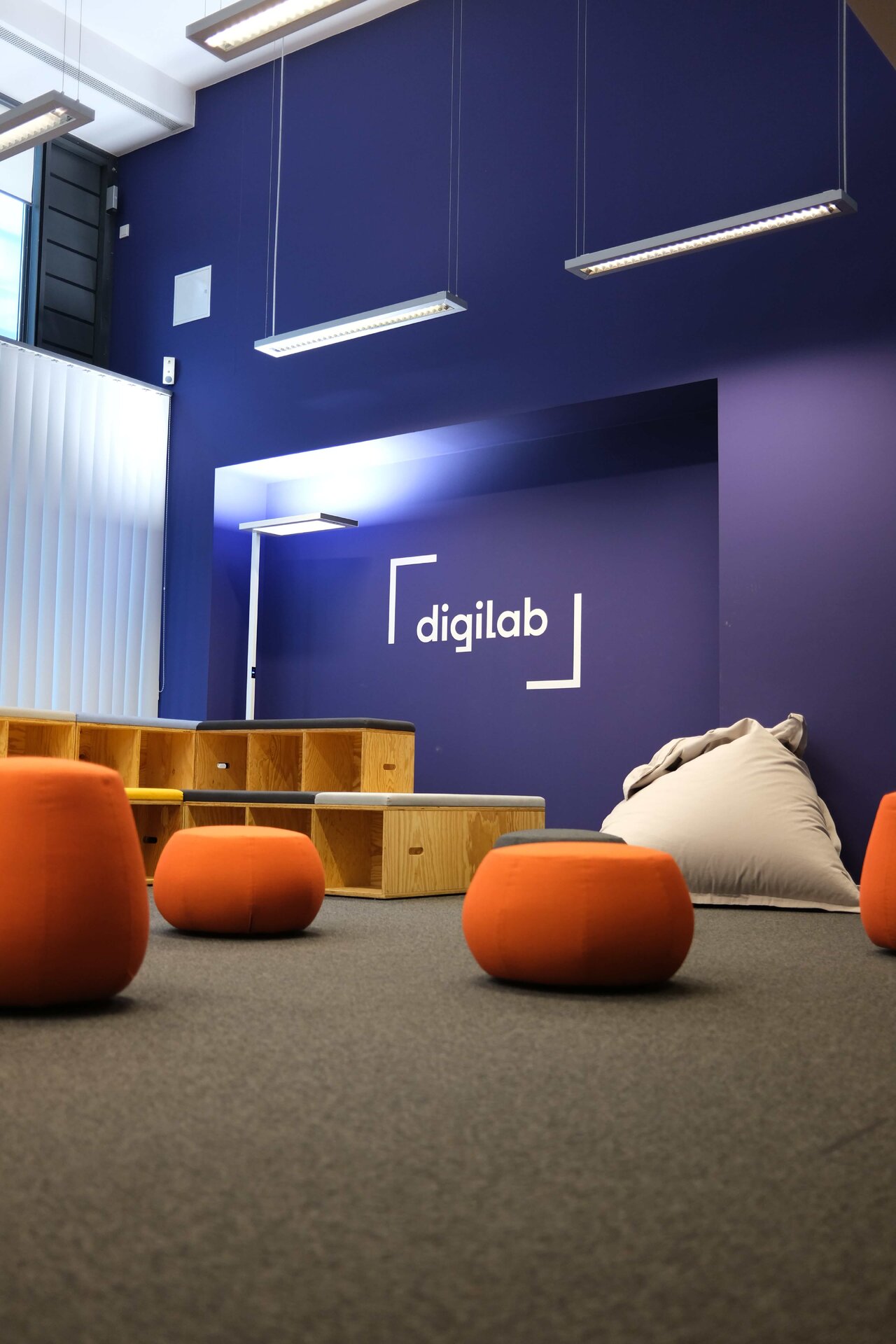 Moderner Gemeinschaftsraum mit blau-violetter Wand und "digilab"-Logo, ausgestattet mit orangefarbenen runden Sitzm&ouml;beln und einem beigen Sitzsack auf grauem Teppichboden, beleuchtet durch lange Deckenleuchten.