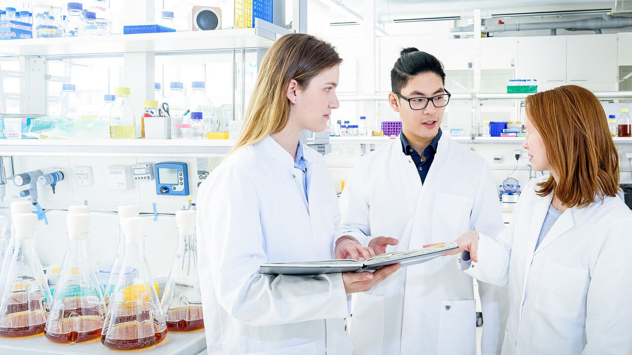 Drei Wissenschaftler in wei&szlig;en Kitteln in einem modernen Laboratorium, umgeben von Glaskolben mit farbigen Fl&uuml;ssigkeiten und wissenschaftlichen Ger&auml;ten, diskutieren und arbeiten zusammen.