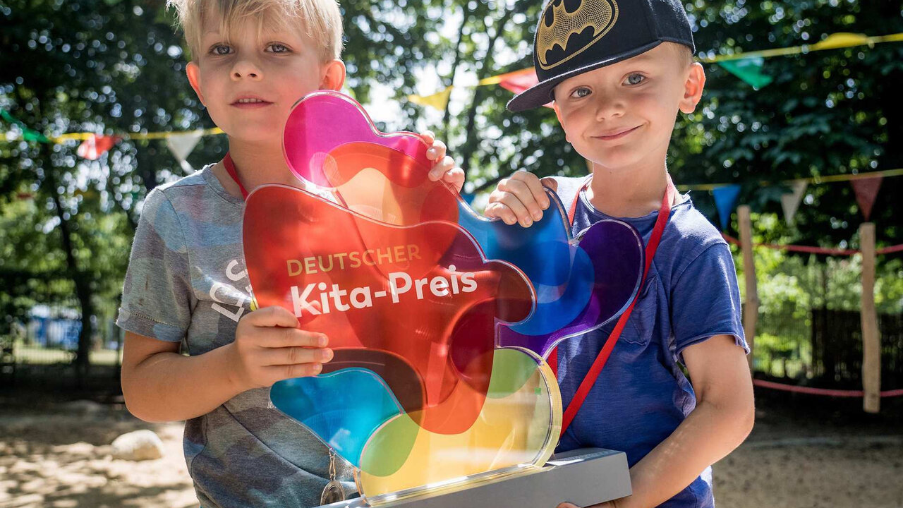 Zwei Jungen halten gemeinsam die mehrfarbige Troph&auml;e des Deutschen Kita-Preises. Sie stehen vor einem unscharfen, gr&uuml;nen Hintergrund und l&auml;cheln selbstbewusst in die Kamera.