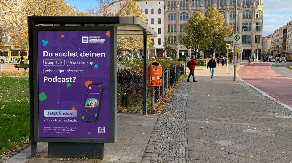 Werbeplakat f&uuml;r Podcast-Suche an einer Bushaltestelle in Berlin, mit lila-farbenem Design und QR-Code, im Hintergrund Gehweg, B&auml;ume und Stadtgeb&auml;ude.