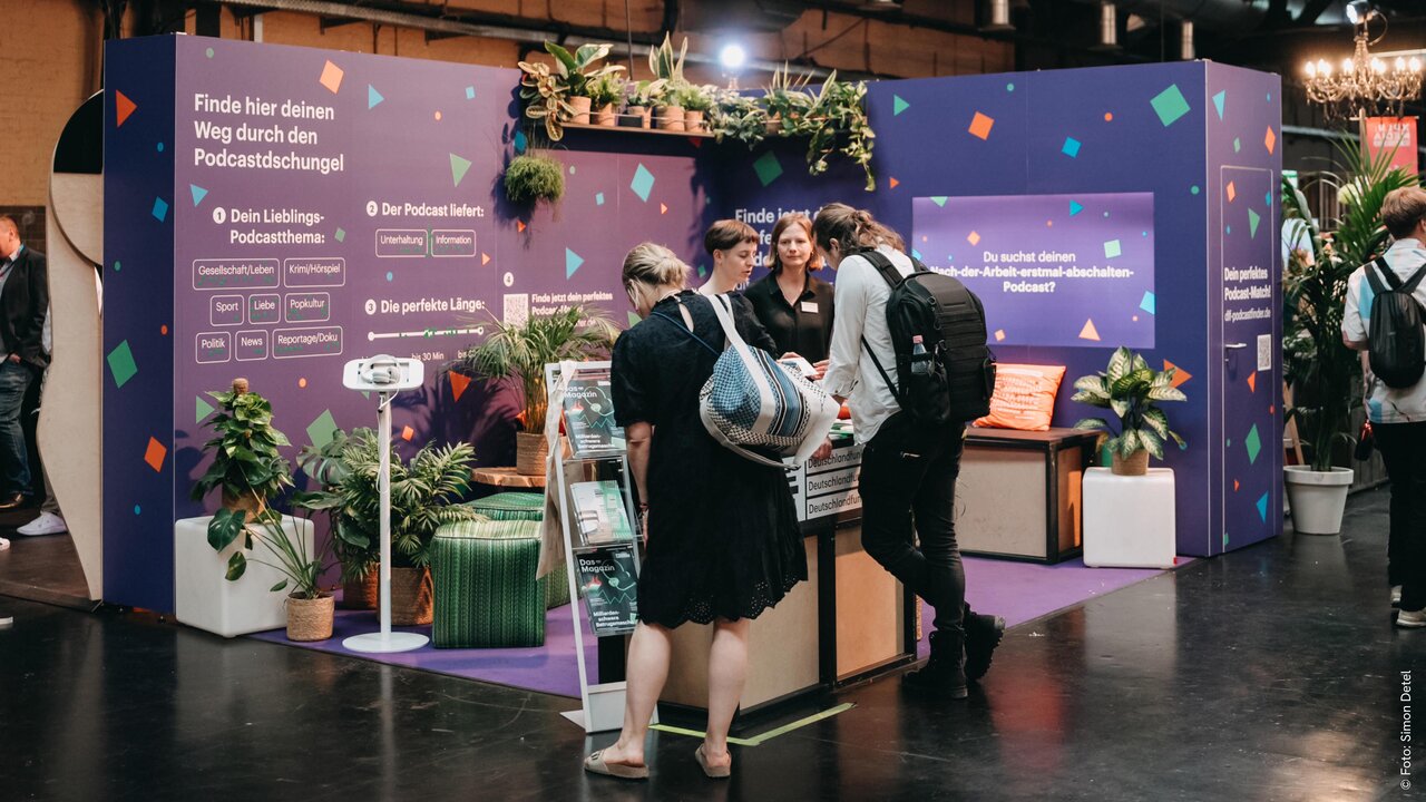 Zwei Personen stehen am Stand einer Veranstaltung mit lila Hintergrund, umgeben von Pflanzen und bunten Konfetti-Elementen, und unterhalten sich in einem professionellen Messeumfeld.