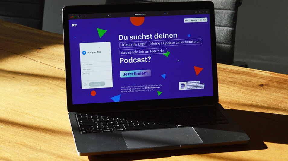 Laptop auf einem Holztisch mit einer Website f&uuml;r Podcast-Suche in blau-violetter Farbgestaltung. Suchformular mit dem Text "Du suchst deinen Podcast?" und bunten grafischen Elementen im Hintergrund.