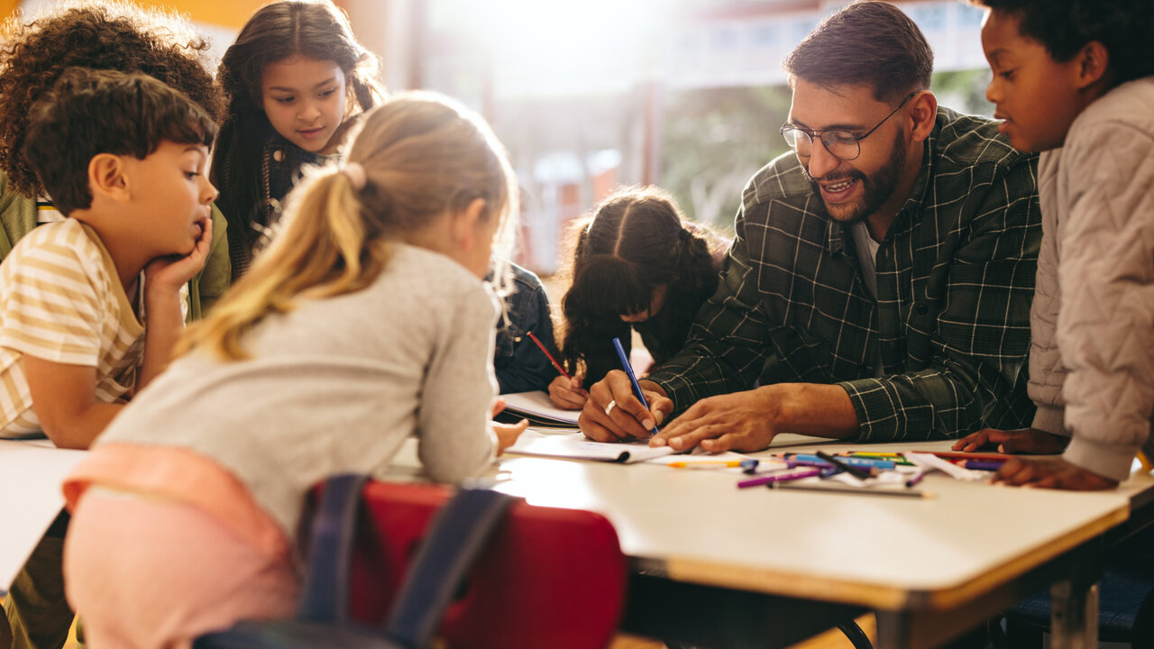 P&auml;dagogische Fachkraft und Kinder sitzen an einem Tisch und arbeiten gemeinsam mit Stiften und Papier. Die Atmosph&auml;re ist konzentriert und freundlich, mit Fokus auf Zusammenarbeit und kreative Bildung.