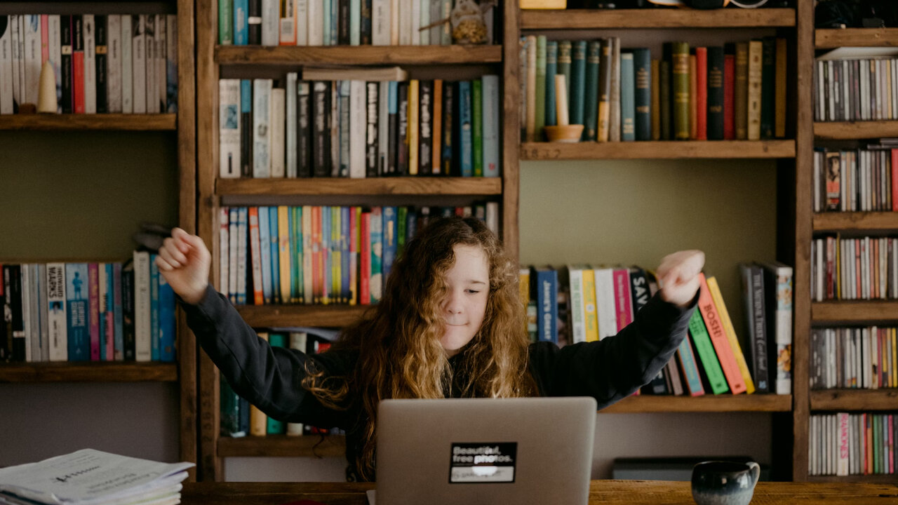 Eine Person mit lockigem Haar sitzt an einem Laptop vor einem B&uuml;cherregal, die Arme ausgestreckt in einer Bewegung der Freude oder Erleichterung. Die Umgebung ist eine gem&uuml;tliche Bibliothek oder ein Arbeitszimmer mit dicht gef&uuml;llten Holzregalen im Hintergrund.