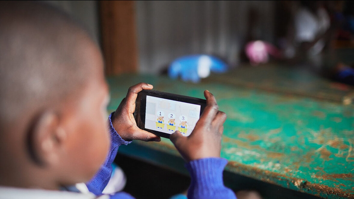 Ein Kind in blauem Pullover sitzt an einem Tisch und spielt auf einem Smartphone mit einer Lern-App, die bunte Icons oder Symbole zeigt.