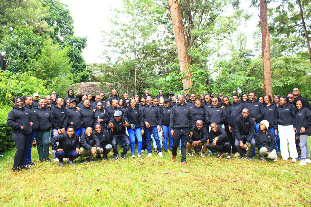 Eine gro&szlig;e Gruppe von Menschen in einheitlichen dunklen Jacken posiert f&uuml;r ein Gruppenfoto auf einer Wiese mit B&auml;umen im Hintergrund. Die Teilnehmer tragen haupts&auml;chlich schwarze Oberbekleidung, mit einigen in blauer und wei&szlig;er Kleidung. Sie stehen und knien eng beieinander, was auf ein Team oder eine Organisationsgruppe hindeutet.