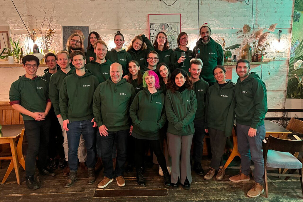 Ein Gruppenfoto des Studio Good Teams in gr&uuml;nen Hoodies, die in einem Raum mit Holzm&ouml;beln und Pflanzen zusammenstehen. Die Gruppe zeigt eine vielf&auml;ltige Mischung von Personen, die freundlich in die Kamera blicken und eine einladende Teamatmosph&auml;re vermitteln.
