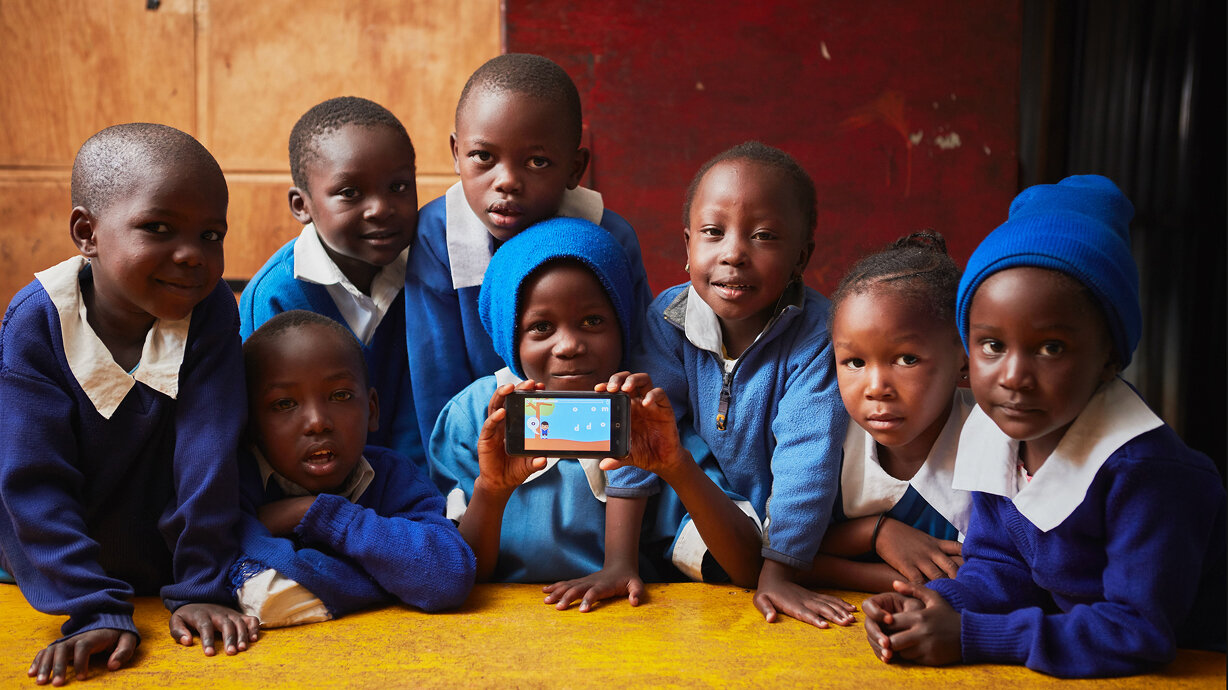 Sieben Schulkinder in blauen Uniformen gruppieren sich um ein Smartphone und blicken fr&ouml;hlich in die Kamera, w&auml;hrend eines der Kinder das Ger&auml;t in der Hand h&auml;lt.