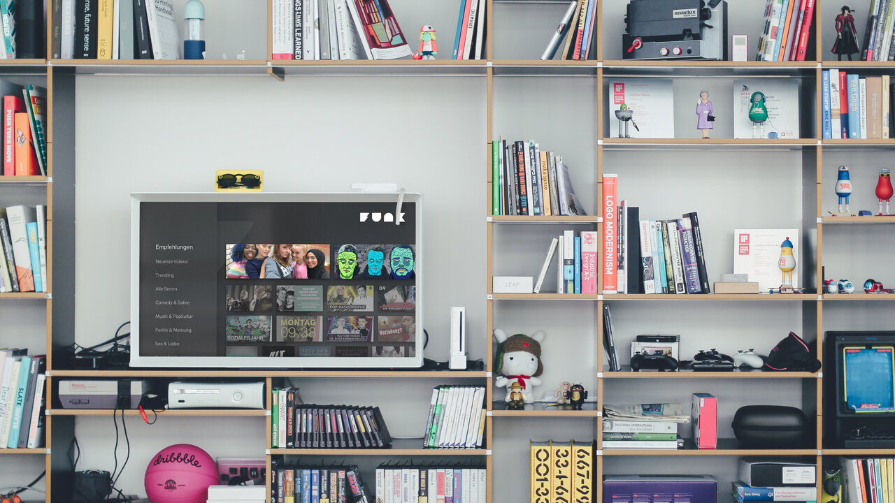 B&uuml;cherregal mit einem Fernseher, der eine AndroidTV App von funk zeigt, mit B&uuml;chern, Dekoration und technischen Ger&auml;ten in einem minimalistischen, modernen Heimarbeitsbereich.