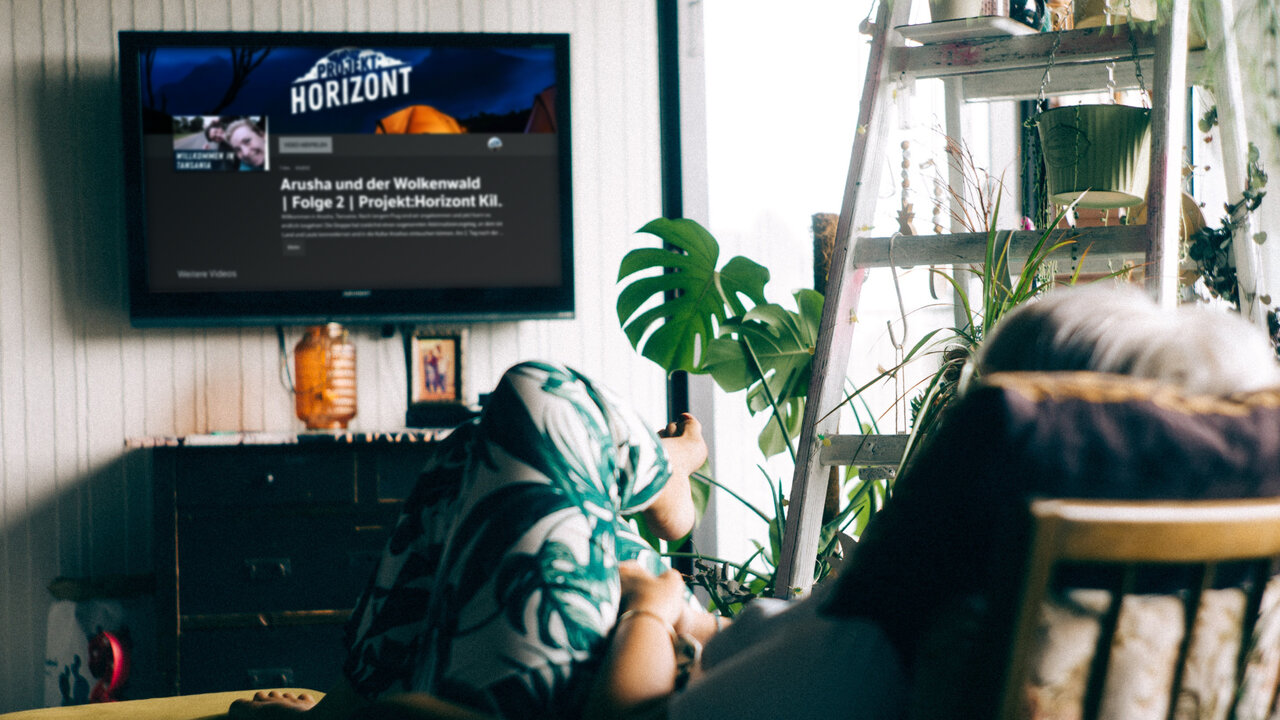 Eine Person sitzt entspannt auf einem Sofa und schaut auf einen Fernseher mit der Horizontapp, umgeben von Zimmerpflanzen und einer Leiter im Hintergrund.