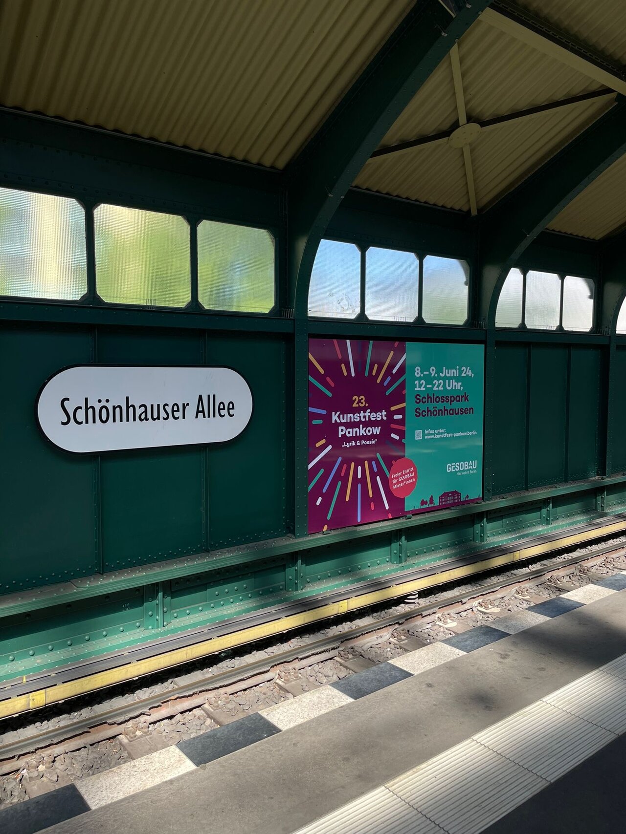 U-Bahnhof Sch&ouml;nhauser Allee mit gr&uuml;n-wei&szlig;er Bahnhofsarchitektur, einem Bahnhofsschild und einem lila-roten Plakat f&uuml;r das Kunstfest Pankow, das Tageslicht f&auml;llt durch die Fenster.