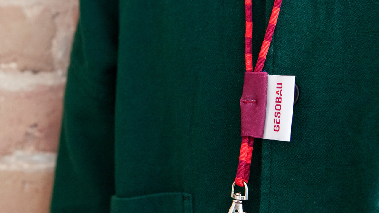 Eine Person mit einer dunkelgr&uuml;nen Jacke, die an eine Backsteinwand gelehnt ist, tr&auml;gt einen roten Lanyard mit GESOBAU-Etikett an.