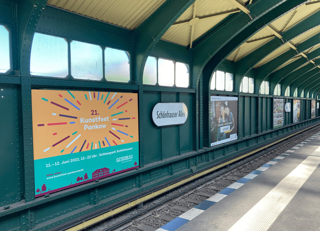 U-Bahnhof mit gr&uuml;n-gelber Konstruktion und einer Plakatwand, die eine Ank&uuml;ndigung f&uuml;r das "21. Kunstfest Pankow" zeigt. Die Station wirkt ruhig und leer, mit klaren Linien und einer strukturierten Architektur.