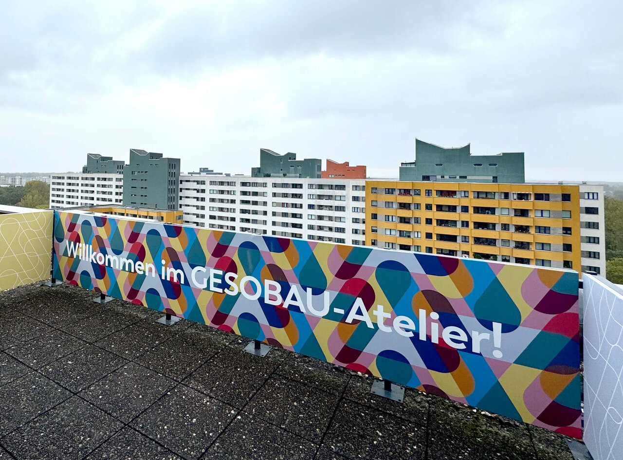 Bunte, abstrakte Wandgestaltung mit dem Text "Willkommen im GESOBAU-Atelier!" vor einem Hintergrund von grauen und gelben Wohnh&auml;usern in Berlin. Die Wandmalerei zeigt ein lebendiges, geometrisches Muster in verschiedenen Farben.