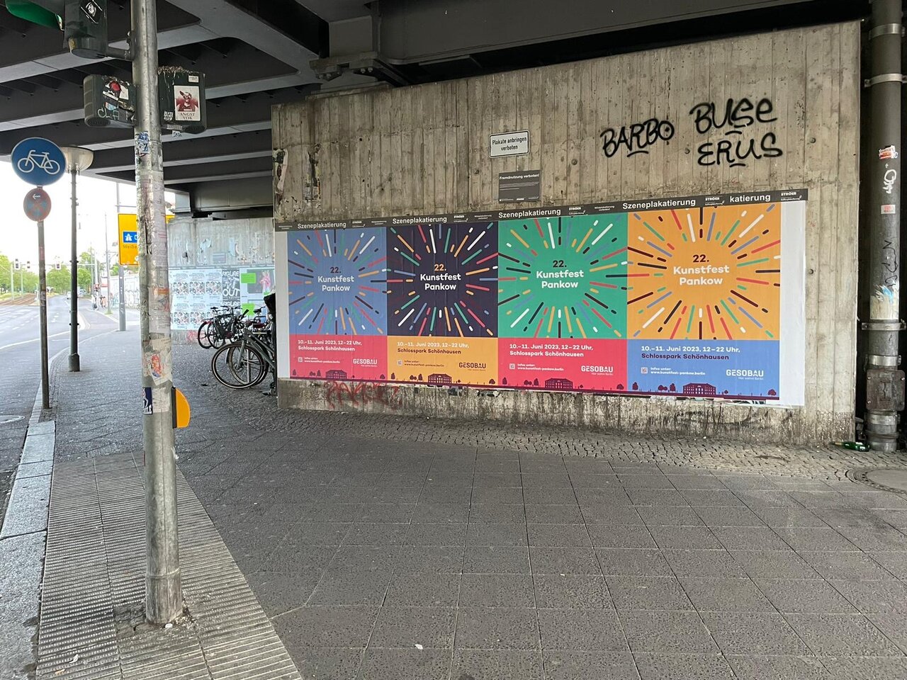 Vier farbenfrohe Plakate mit Strahlenmotiven an einer Betonwand unter einer Br&uuml;cke, die verschiedene Botschaften in unterschiedlichen Farben pr&auml;sentieren. Fahrr&auml;der und Verkehrszeichen sind im Hintergrund zu sehen.