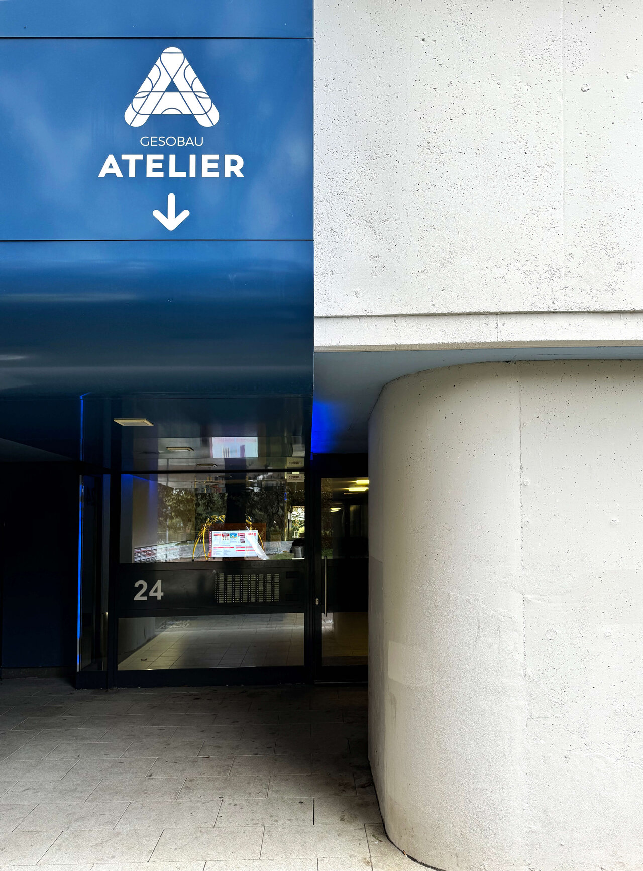 Blauer Geb&auml;udeeingang mit GESOBAU Atelier Schild, Glasfront mit Hausnummer 24 und geschwungener wei&szlig;er Wand, moderner Architekturkontrast in urbaner Umgebung.
